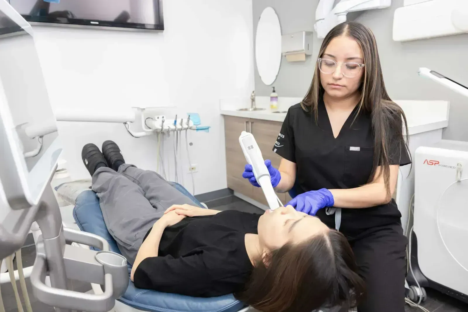 A dental hygienist in black scrubs examines a patient reclined in a dental chair.
